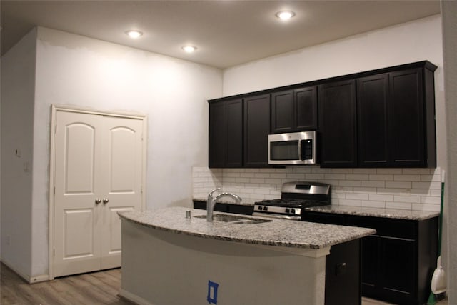 kitchen with sink, a kitchen island with sink, light stone counters, stainless steel appliances, and light hardwood / wood-style flooring