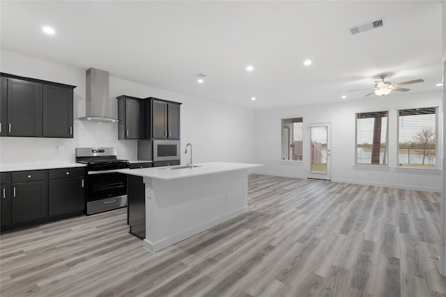 kitchen featuring sink, stainless steel gas range oven, an island with sink, light hardwood / wood-style floors, and wall chimney range hood