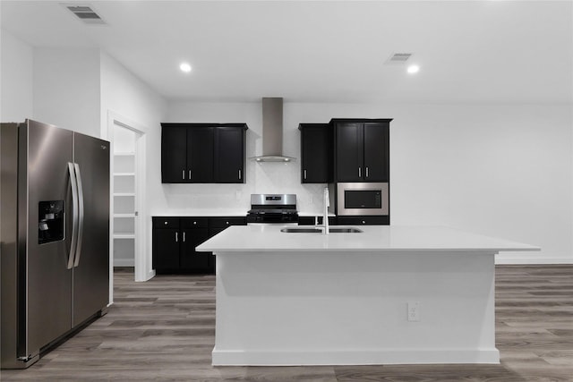 kitchen with wall chimney range hood, sink, stainless steel appliances, and an island with sink