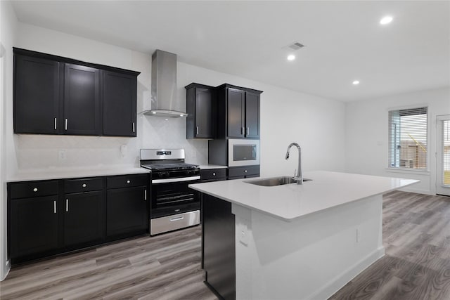 kitchen with sink, wall chimney range hood, a center island with sink, stainless steel gas range, and light wood-type flooring