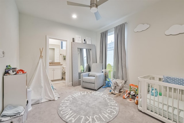 bedroom with a crib, connected bathroom, light colored carpet, and ceiling fan