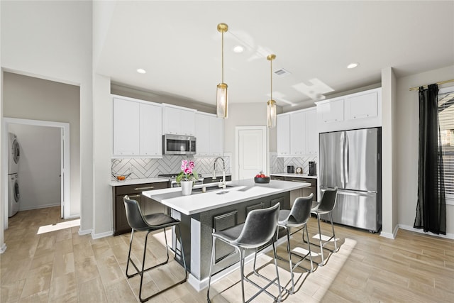 kitchen featuring appliances with stainless steel finishes, an island with sink, white cabinetry, hanging light fixtures, and stacked washer and clothes dryer