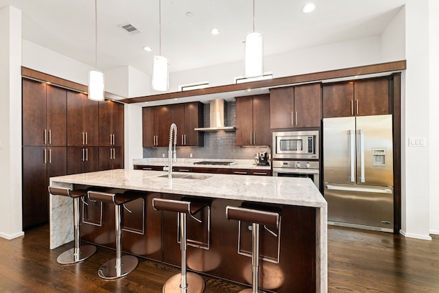 kitchen with a center island with sink, hanging light fixtures, built in appliances, and wall chimney exhaust hood