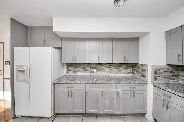 kitchen featuring backsplash, gray cabinetry, light stone counters, and white fridge with ice dispenser