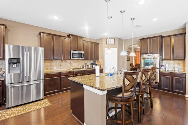 kitchen with sink, hanging light fixtures, an island with sink, stainless steel appliances, and light stone countertops