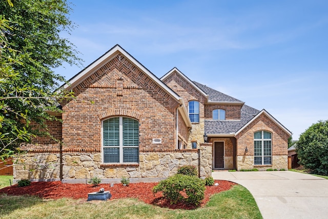 french country home featuring driveway, brick siding, roof with shingles, and stone siding