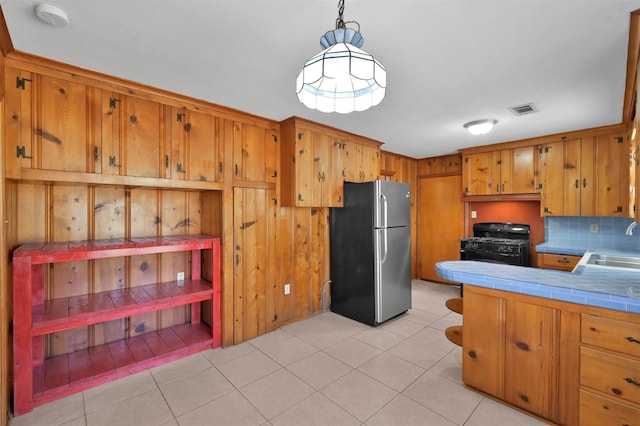 kitchen with stainless steel refrigerator, sink, hanging light fixtures, tile counters, and gas range oven