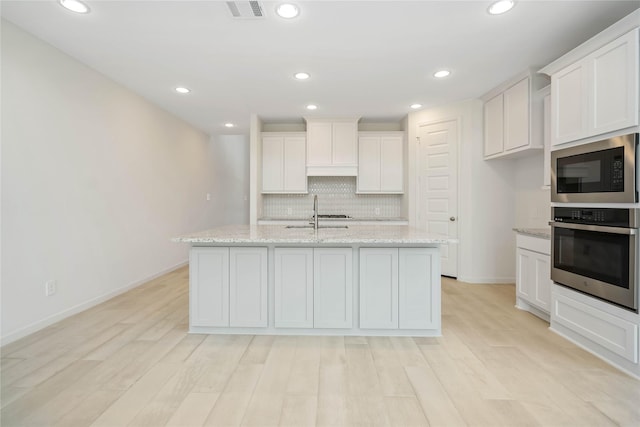 kitchen with light stone counters, white cabinets, a center island with sink, built in microwave, and oven