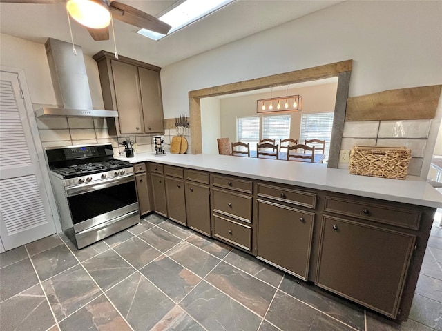 kitchen featuring backsplash, dark brown cabinetry, kitchen peninsula, wall chimney range hood, and stainless steel gas range