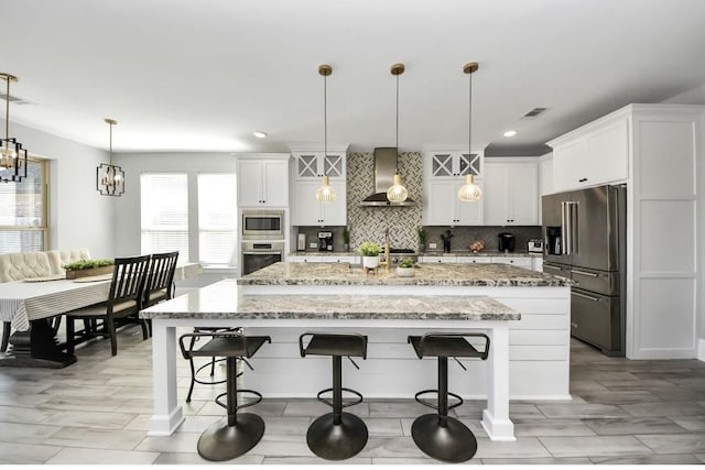 kitchen with white cabinetry, stainless steel appliances, an island with sink, decorative light fixtures, and wall chimney exhaust hood