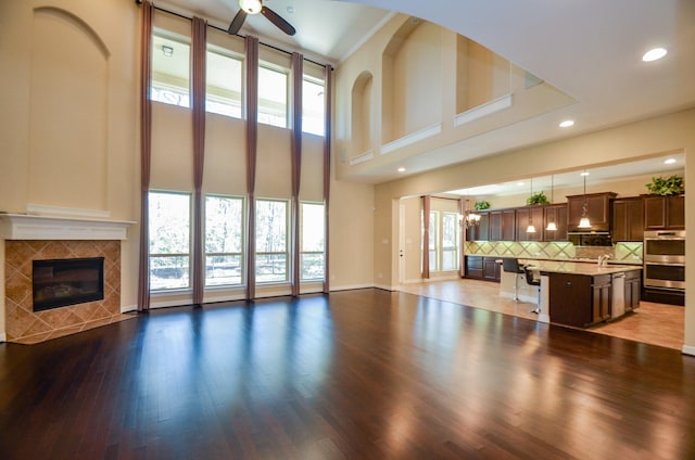 unfurnished living room with a towering ceiling, ceiling fan with notable chandelier, sink, a tiled fireplace, and hardwood / wood-style flooring