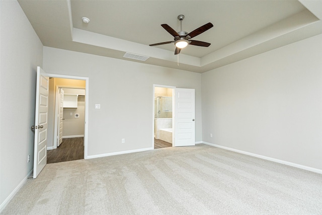 unfurnished bedroom with ceiling fan, ensuite bathroom, carpet flooring, a tray ceiling, and a spacious closet