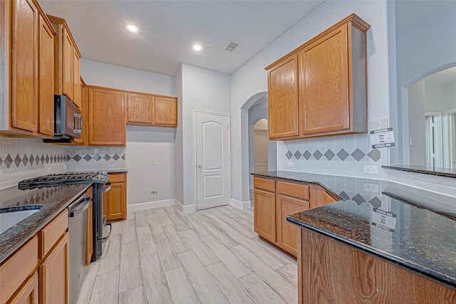 kitchen featuring appliances with stainless steel finishes, light hardwood / wood-style floors, dark stone counters, and backsplash