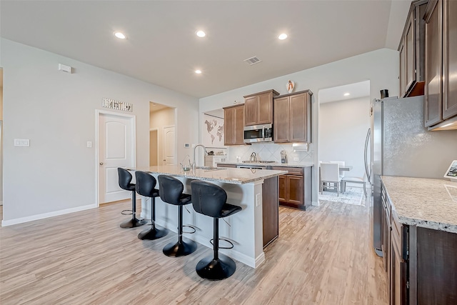 kitchen featuring sink, a kitchen island with sink, stainless steel appliances, light stone countertops, and light wood-type flooring