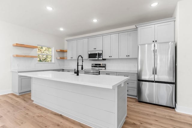 kitchen featuring appliances with stainless steel finishes, tasteful backsplash, gray cabinetry, a kitchen island with sink, and light hardwood / wood-style flooring