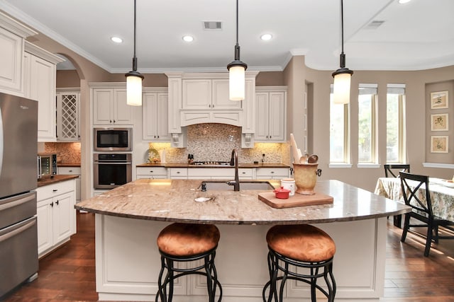 kitchen featuring stainless steel appliances, an island with sink, light stone counters, and decorative light fixtures