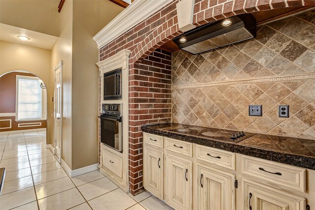 kitchen featuring tasteful backsplash, black appliances, light tile patterned flooring, cream cabinetry, and wall chimney exhaust hood