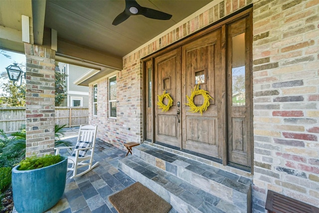 view of exterior entry with brick siding, a porch, ceiling fan, and fence