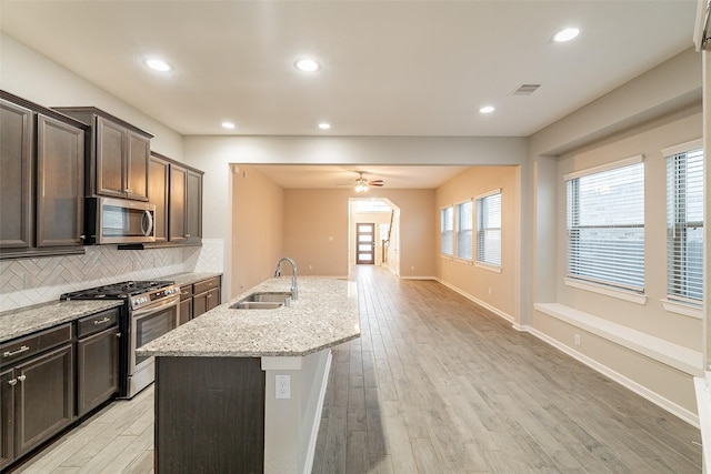 kitchen featuring sink, backsplash, stainless steel appliances, light stone countertops, and an island with sink