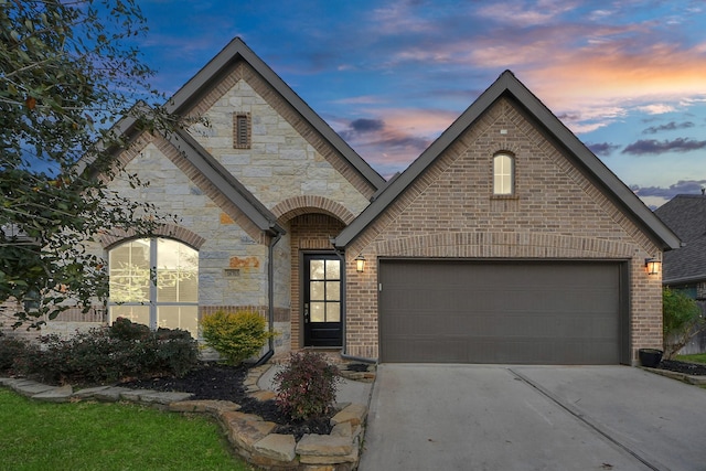 french country inspired facade featuring a garage, stone siding, concrete driveway, and brick siding