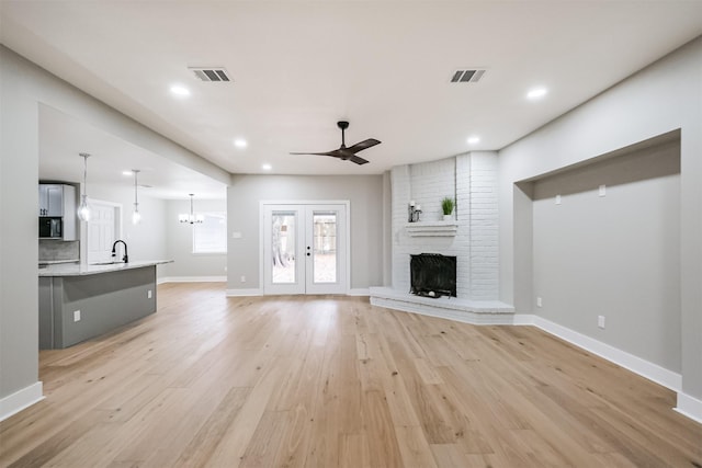 unfurnished living room with french doors, sink, a brick fireplace, light hardwood / wood-style floors, and ceiling fan with notable chandelier