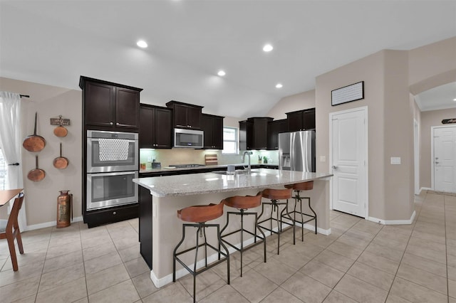 kitchen featuring a kitchen bar, a center island with sink, light tile patterned floors, stainless steel appliances, and light stone countertops