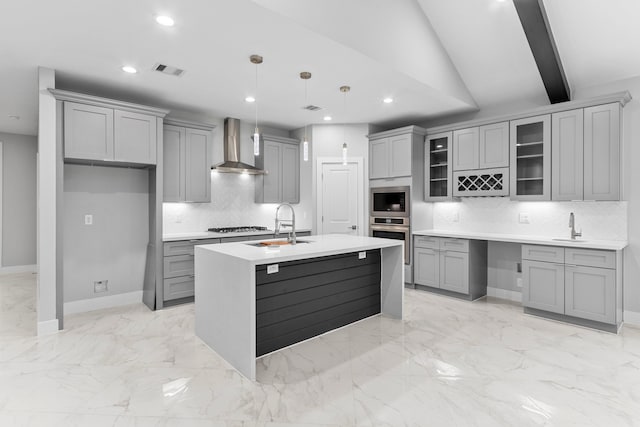 kitchen featuring wall chimney range hood, backsplash, vaulted ceiling with beams, hanging light fixtures, and an island with sink