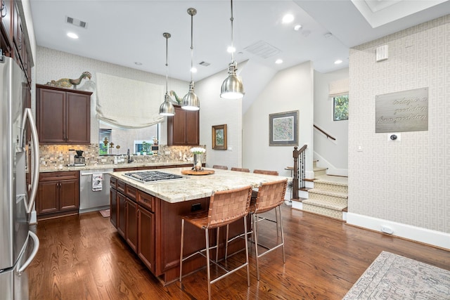 kitchen with dark wood-type flooring, appliances with stainless steel finishes, hanging light fixtures, light stone countertops, and a kitchen island