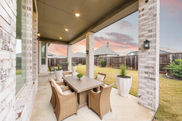 patio terrace at dusk featuring outdoor dining area, a fenced backyard, and a lawn