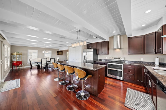 kitchen featuring wall chimney range hood, appliances with stainless steel finishes, beam ceiling, a center island, and decorative light fixtures