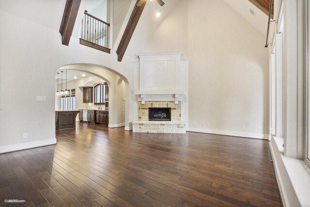 unfurnished living room featuring a stone fireplace, high vaulted ceiling, dark hardwood / wood-style flooring, beamed ceiling, and ceiling fan