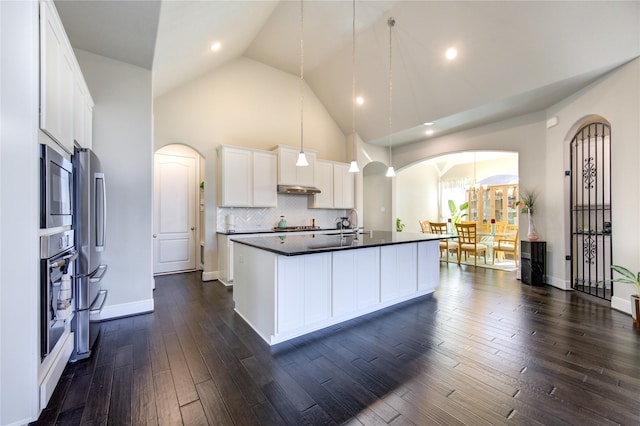 kitchen featuring arched walkways, a center island with sink, dark countertops, appliances with stainless steel finishes, and white cabinets