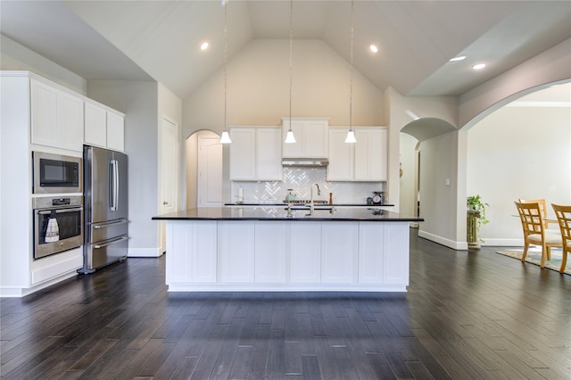 kitchen featuring arched walkways, a kitchen island with sink, stainless steel appliances, white cabinets, and dark countertops