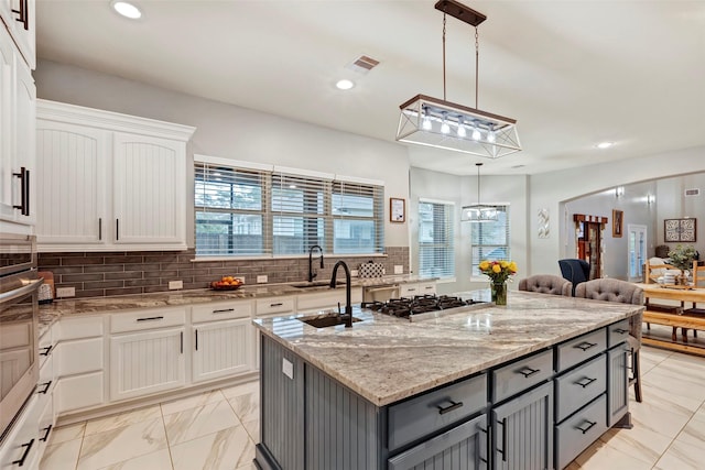 kitchen featuring light stone counters, a center island with sink, gray cabinets, stainless steel gas stovetop, and white cabinets