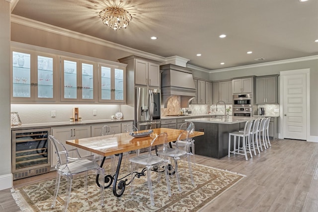 kitchen with wine cooler, premium range hood, appliances with stainless steel finishes, a kitchen breakfast bar, and a kitchen island with sink