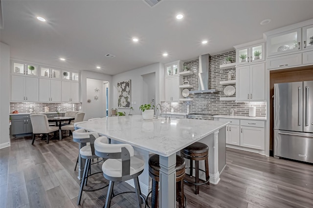 kitchen with wall chimney range hood, high end fridge, light stone countertops, white cabinets, and a center island with sink