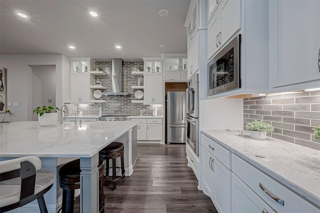 kitchen featuring white cabinetry, a kitchen bar, stainless steel appliances, light stone countertops, and wall chimney range hood