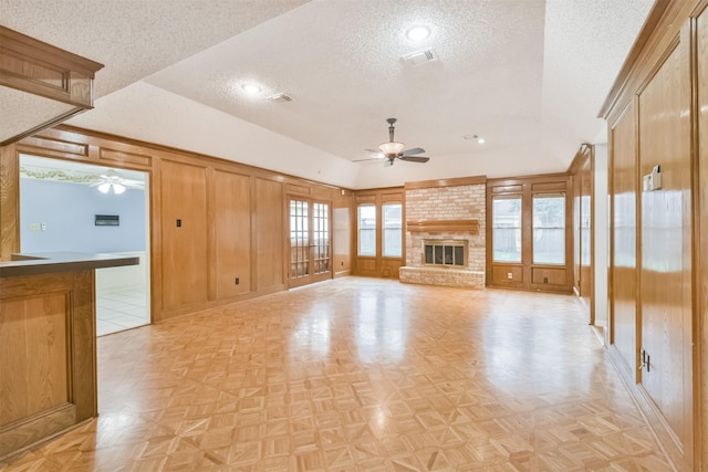 unfurnished living room featuring a brick fireplace, light parquet floors, ceiling fan, and wood walls