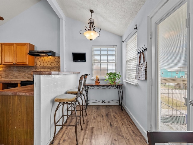 kitchen featuring lofted ceiling, a textured ceiling, under cabinet range hood, brown cabinets, and light wood finished floors