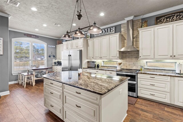 kitchen featuring pendant lighting, wall chimney range hood, stainless steel appliances, a kitchen island, and dark stone counters