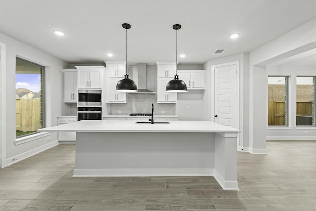 kitchen featuring stainless steel microwave, white cabinetry, oven, wall chimney range hood, and a center island with sink