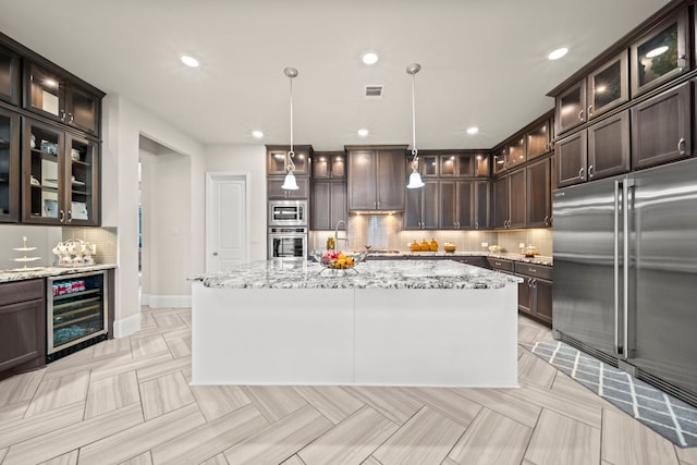 kitchen with pendant lighting, built in appliances, light stone countertops, a center island with sink, and beverage cooler