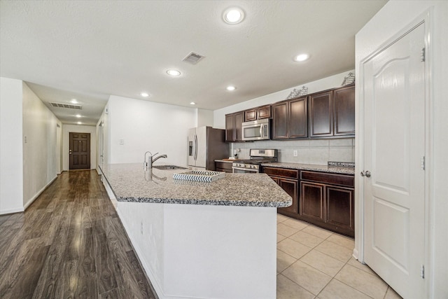 kitchen with sink, dark brown cabinetry, stainless steel appliances, light stone countertops, and a center island with sink