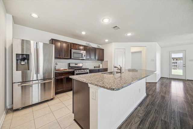 kitchen with appliances with stainless steel finishes, sink, backsplash, a kitchen island with sink, and light stone counters