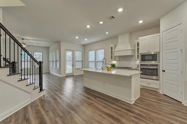 kitchen with stainless steel appliances, an island with sink, dark hardwood / wood-style flooring, decorative backsplash, and custom exhaust hood