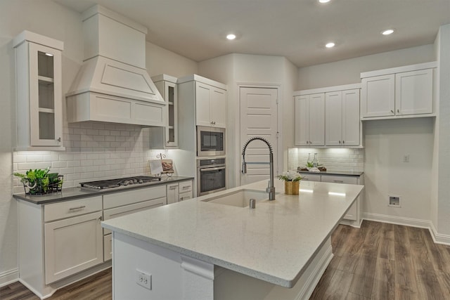 kitchen with dark wood-style flooring, a center island with sink, stainless steel appliances, a sink, and light stone countertops