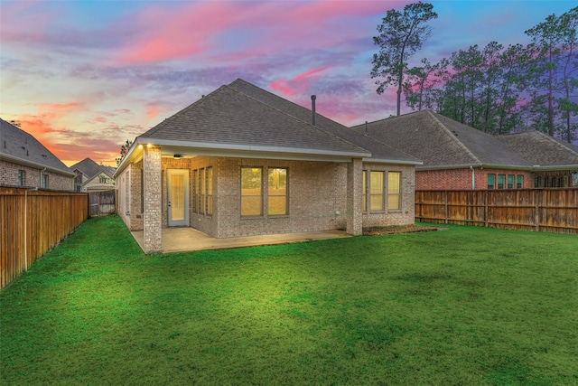 back of property at dusk with a fenced backyard, brick siding, a yard, roof with shingles, and a patio area