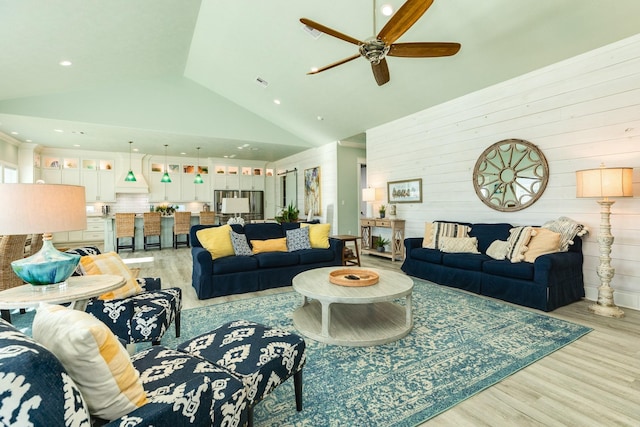 living room featuring high vaulted ceiling, light hardwood / wood-style floors, ceiling fan, and wood walls