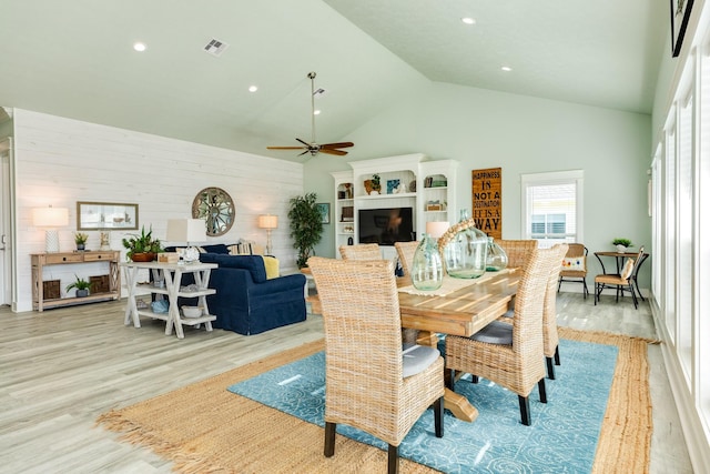 dining space featuring ceiling fan, high vaulted ceiling, light wood-type flooring, and wood walls