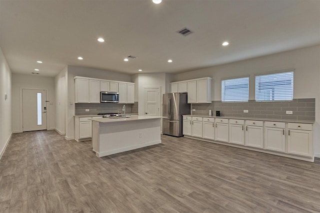 kitchen featuring stainless steel appliances, tasteful backsplash, white cabinets, a center island with sink, and light wood-type flooring